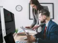 Para omawia dokumenty przy biurku, analizując dane. W tle zegar i monitory. To ich codzienna praca, być może związana z zus.pl.