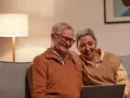 Uśmiechnięci seniorzy oglądają coś na laptopie. Mężczyzna, pracujący emeryt na zwolnieniu lekarskim, śmieje się, a kobieta opiera rękę na jego ramieniu.