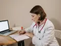 Lekarka w białym kitlu z czerwonym stetoskopem siedzi przy biurku, pisząc w zeszycie. Obok leży laptop. Czy sor wystawia L4?