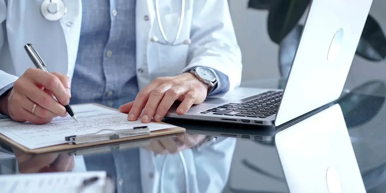 Lekarz w białym kitlu pisze na dokumentach, przygotowując zwolnienie lekarskie ze szpitala. Obok leży laptop.
