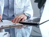 Lekarz w białym kitlu pisze na dokumentach, przygotowując zwolnienie lekarskie ze szpitala. Obok leży laptop.