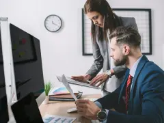 Para omawia dokumenty przy biurku, analizując dane. W tle zegar i monitory. To ich codzienna praca, być może związana z zus.pl.