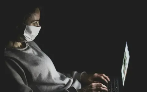 Kobieta w masce medycznej pracuje na laptopie w ciemności.