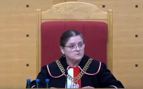 Sędzia w todze i łańcuchu na szyi, siedzi za stołem z butelkami wody. Czy sędziowie płacą składkę zdrowotną?