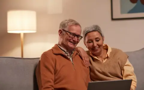 Uśmiechnięci seniorzy oglądają coś na laptopie. Mężczyzna, pracujący emeryt na zwolnieniu lekarskim, śmieje się, a kobieta opiera rękę na jego ramieniu.
