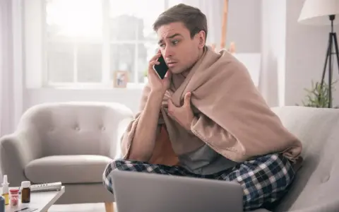 Chory mężczyzna owinięty kocem rozmawia przez telefon, martwiąc się, czy sobota i niedziela wlicza się do chorobowego.