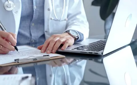 Lekarz w białym kitlu pisze na dokumentach, obok laptop.