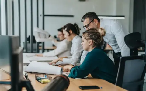 Zespół pracuje nad projektem. Mężczyzna pochyla się nad kobietą, wyjaśniając coś. To przykład, co to jest pracownik najemny.