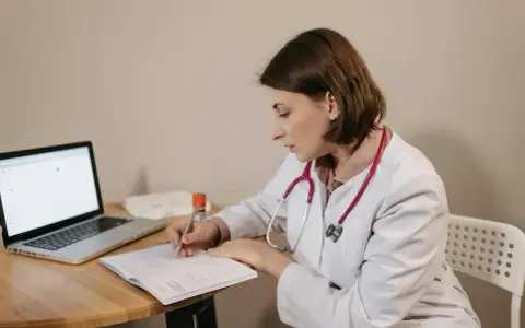 Lekarka w białym kitlu z czerwonym stetoskopem siedzi przy biurku, pisząc w zeszycie. Obok leży laptop. Czy sor wystawia L4?