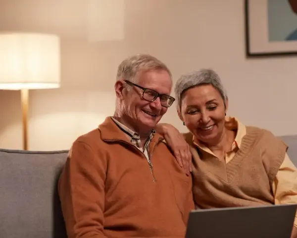 Uśmiechnięci seniorzy oglądają coś na laptopie. Mężczyzna, pracujący emeryt na zwolnieniu lekarskim, śmieje się, a kobieta opiera rękę na jego ramieniu.