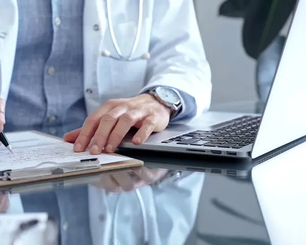 Lekarz w białym kitlu pisze na dokumentach, obok laptop.