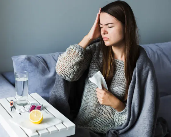 Kobieta z bólem głowy, okryta kocem, z chusteczką w dłoni. Na stoliku leki, cytryna i woda. Co dalej, gdy przerwane zwolnienie lekarskie przez orzecznika ZUS?