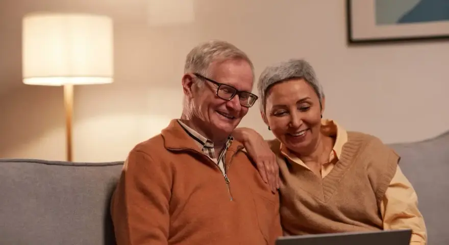 Uśmiechnięci seniorzy oglądają coś na laptopie. Mężczyzna, pracujący emeryt na zwolnieniu lekarskim, śmieje się, a kobieta opiera rękę na jego ramieniu.