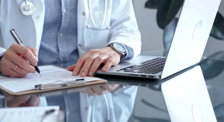 Lekarz w białym kitlu pisze na dokumentach, obok laptop.