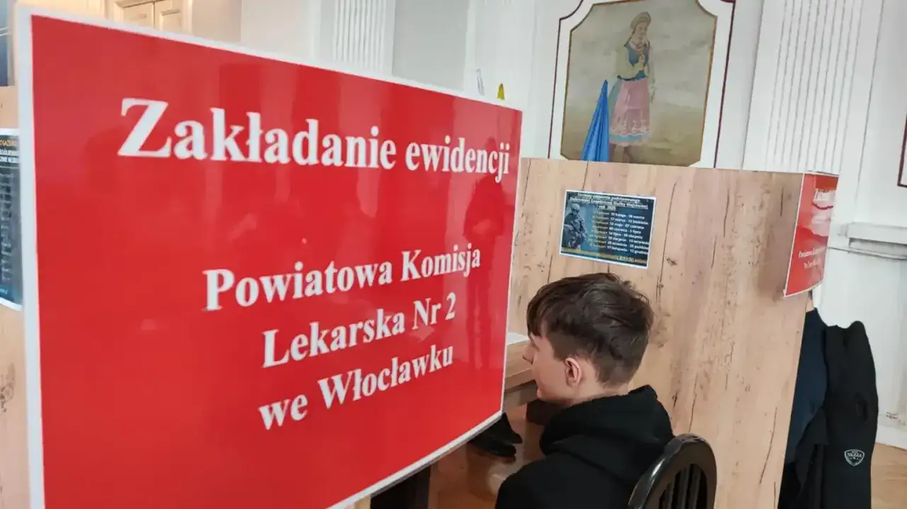 Młody mężczyzna czeka na badanie na komisji wojskowej. Na czerwonej tablicy widnieje napis 